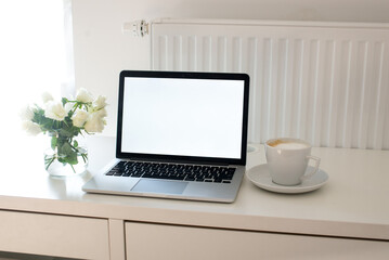 Fototapeta premium Laptop with white blank screen on white work table. White interior home office concept. Female office desk workspace with white roses and cup of coffee. Close up on screen laptop. Mock up laptop
