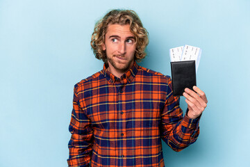 Young caucasian man holding a passport isolated on blue background confused, feels doubtful and unsure.