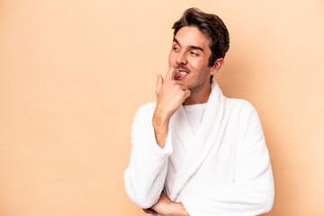 Young caucasian man wearing a bathrobe isolated on beige background relaxed thinking about something looking at a copy space.