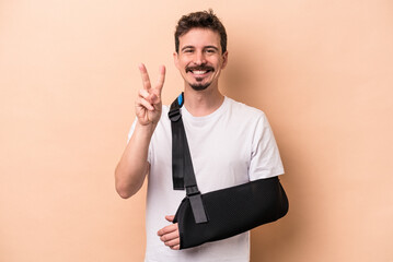 Young caucasian man with broken arm isolated on beige background showing number two with fingers.