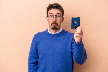 Young informatic man isolated on beige background shrugs shoulders and open eyes confused.