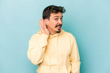 Young caucasian man isolated on blue background trying to listening a gossip.