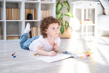 curly child girl drawing with colored markers lying on floor in living room at home