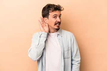 Young caucasian man isolated on beige background trying to listening a gossip.