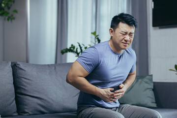 Asian man himself at home sick sitting on the couch, has severe abdominal pain, man in the living...