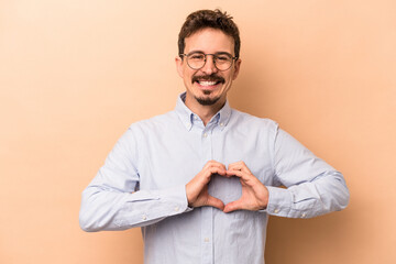 Young caucasian man isolated on beige background smiling and showing a heart shape with hands.