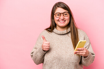 Young caucasian overweight woman holding a mobile phone isolated on pink background smiling and raising thumb up