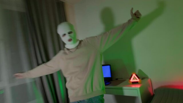 A masked gangster looks menacingly at the camera. Frame for a rap video. A green neon machine illuminates the room. 4k video at long exposure.