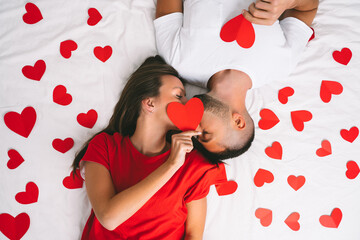 Top view of beautiful couple in love lying on white bed with red hearts and kissing. Love and relationships concept