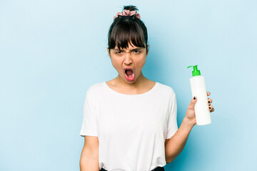 Young hispanic woman holding a body lotion isolated on blue background screaming very angry and...