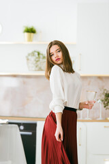 Portrait of fashionable woman in a red skirt and white blouse posing on the kitchen. Girl with a vine glass in a hands