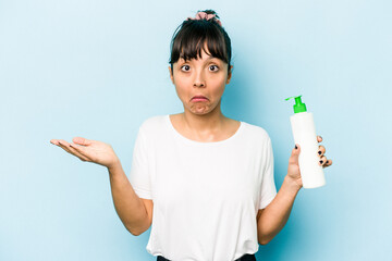 Young hispanic woman holding a body lotion isolated on blue background shrugs shoulders and open eyes confused.