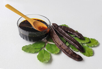 carob molasses in glass bowl and in wooden spoon and carob pods on rustic background, locust bean healthy food, Ceratonia siliqua ( harnup )