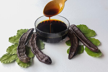 carob molasses in glass bowl and in wooden spoon and carob pods on rustic background, locust bean healthy food, Ceratonia siliqua ( harnup ) - obrazy, fototapety, plakaty