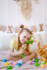 easter, a little girl with painted colored eggs and a rabbit holding spring flowers tulips at home in a bright room preparing for the holiday smiling and having fun