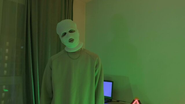 A masked gangster looks menacingly at the camera. Frame for a rap video. A green neon machine illuminates the room.