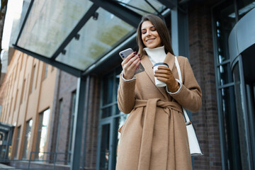 Cheerful young woman wearing brown coat standing in downtown holding paper coffee cup and using mobile phone