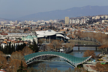 Naklejka premium Aerial view of the Bridge of Peace, a pedestrian bridge made of steel and glass with a curvy design, over the Kura Mtkvari river in Tbilisi, Georgia. The Public Service Hall is seen in the background.