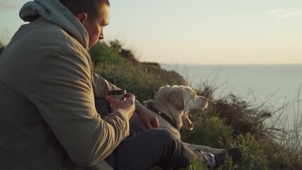 Young handsome male caress his retriever dog sitting on a cliff near sea