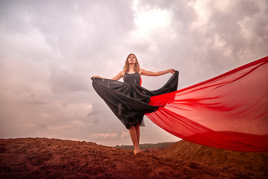 Young slim girl in black dress with red fabric dances on sand dunes against a dramatic sky before a thunderstorm. Model posing during photo shoot on nature