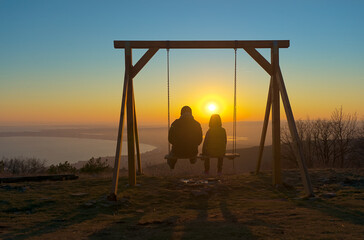 A man and a child are sitting on a swing on a hill overlooking a beautiful landscape at sunset, the sea and the valley