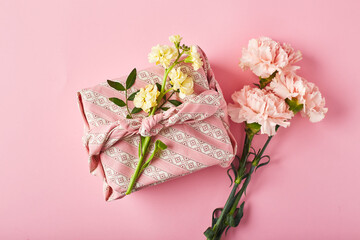 Obraz na płótnie Canvas Bouquet of pink carnations and yellow matthiola with gift wrap in traditional japanese furoshiki style.. Design concept of holiday greeting with carnation bouquet on pink table background