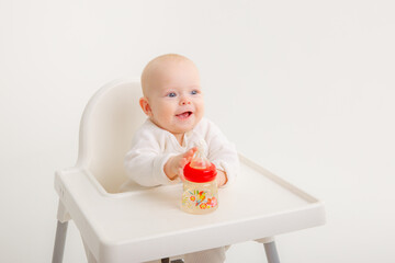 little baby  drinks water from a bottle indoors, sits on a high chair