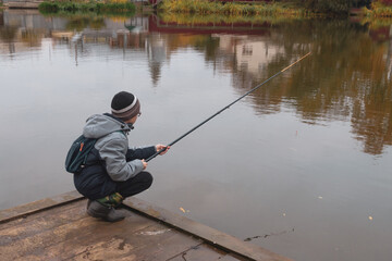 A teenage Caucasian boy is fishing on a calm autumn day on the lake squatting