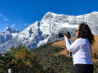 Woman with curly hair shooting on camera of beautiful panorama of snow capped Jade Dragon Snow Mountain with yellow meadows and forest. Located near Shangri - La. Tibet, China