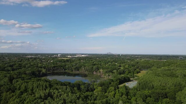 Aerial view over Lake Minnetonka with clear blue sky with wispy clouds. Lush green foliage all around. Water from the lake reflecting the sun.  Park tucked into forest. 