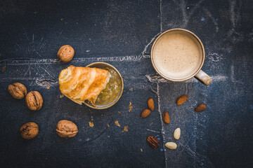 Piece of delicious French croissant dipped in jam, a cup of coffee and some nuts, breakfast scene, black background, top view