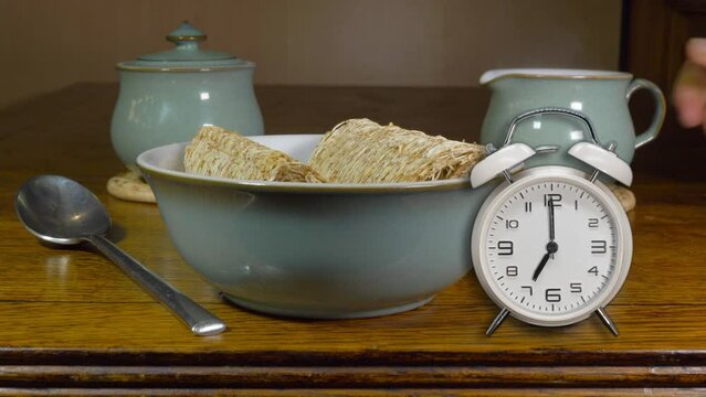 A composite video of dry shredded wheat cereal in a bowl, on a table next to a miniature traditional alarm clock with bells ringing at 7 o’clock, and a hand putting down a jug of steaming hot milk.