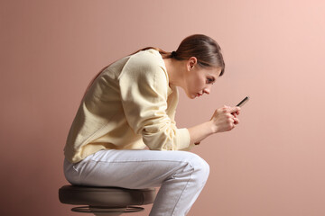 Woman with bad posture using smartphone while sitting on stool against pale pink background