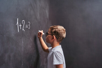 Primary education. Side view student solves math example on a blackboard in math class. an elementary school student stands at blackboard and writes an answer to an example, thinking about the answer.