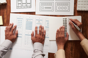 Hands of UI and UX designers working with wireframes and layouts of mobile application, view from above