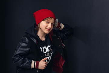 cute pre-teen boy with a nose piercing, in fashionable clothes stands near a black wall and looks into the camera. Adolescence. The concept of youth and clothing.