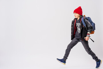 Portrait of teenage schoolboy in fashionable clothes with backpack on his shoulder, full-length, walking on white background. Adolescence. Advertising of teenage clothes and satchels. Copy space