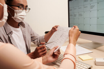 Team of UI designers in medical masks discussing hand drawn wireframe at meeting