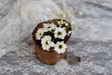 White daisies and baby breath in rusted tin can with feathers on white lace cloth. 