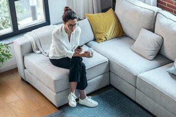 Pretty young woman using mobile phone while drinking cup of coffee on sofa at home.