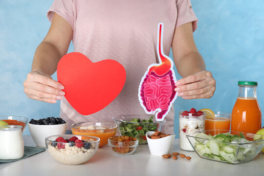 Woman holding paper intestine and heart near table with food, closeup. Balanced nutrition for healthy digestive system
