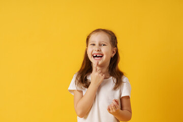 cute smiling preschool girl holds her first fallen baby tooth in her hand while standing on yellow...