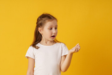surprised preschool girl holds her first dropped baby tooth in her hand while standing on a yellow...