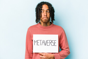 Young African American man holding metaverse placard isolated on blue background confused, feels doubtful and unsure.