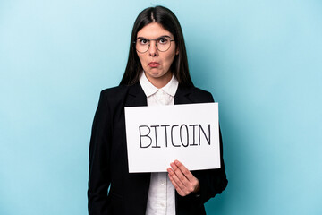 Young caucasian business woman holding a bitcoin placard isolated on blue background shrugs...