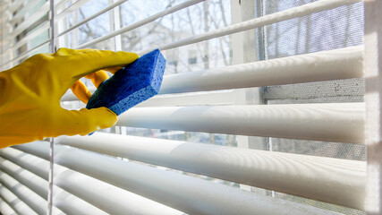 Hand with yellow glove and blue sponge is cleaning aluminum window blinds