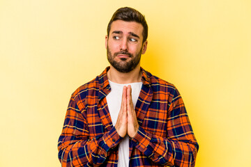 Young caucasian man isolated on yellow background praying, showing devotion, religious person looking for divine inspiration.