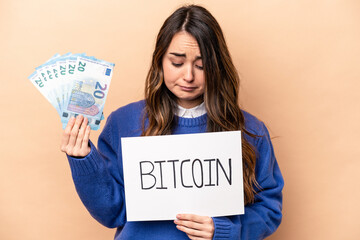 Young caucasian woman holding a bitcoin placard and banknotes isolated on beige background