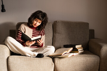 A mis adult Spanish brunette woman is reading a book on her sofa with many books around her in her living room at home, comfy clothes, indoors © Sandra