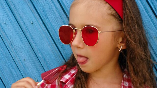 The child licks the red triangular lollipop with pleasure. Curly brunette Girl in sunglasses on a blue background. The harm of Eating sugar and sweets